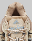 Close-up of the tongue and upper area of an Adidas Pharrell Williams Adistar Jellyfish - "Crystal Sand" sneaker in beige and cream tones, with light blue adidas branding and 'VIRGINIA' text on the lace guard, set against a solid gray background.