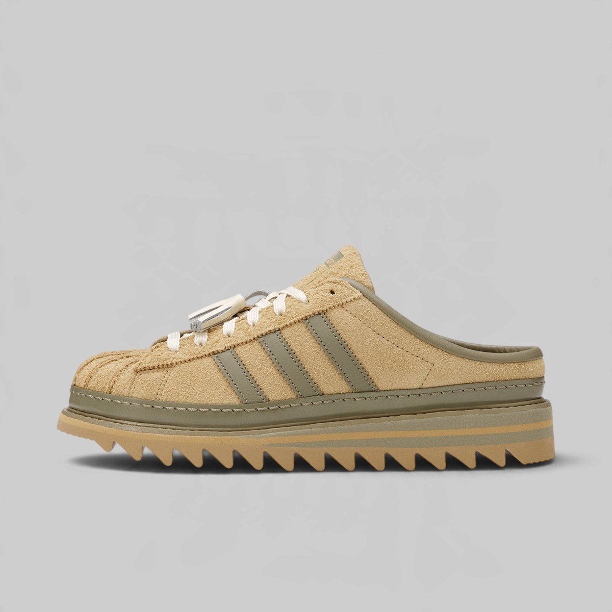A low-top slip-on sneaker in beige suede with olive green stripes and laces, featuring a chunky gum sole with a zigzag tread, shown against a solid gray background.