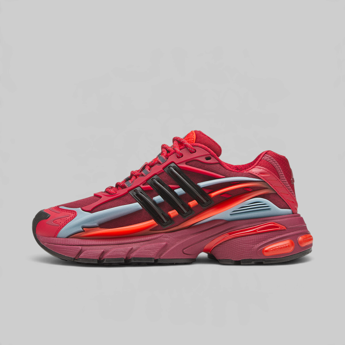 A red Adidas sneaker with black three-stripes, a translucent light blue side panel, and a bright orange midsole accent, shown in profile against a light gray background.