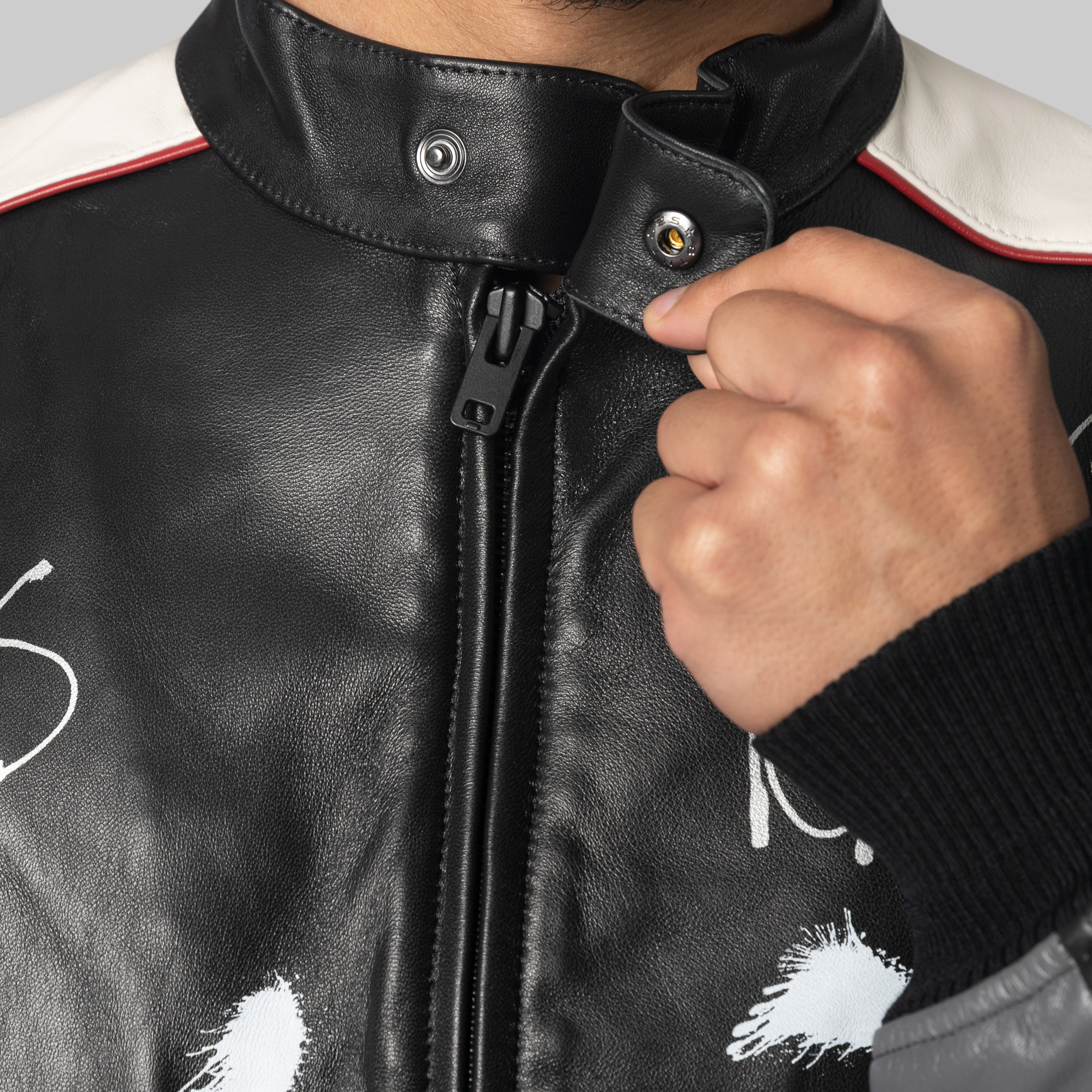 Close-up of a person wearing a black leather motorcycle jacket with a high collar, white shoulder panels, and white paint-splatter graphics, adjusting the zipper with a hand.