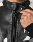 Close-up of a person wearing a black leather motorcycle jacket with a high collar, white shoulder panels, and white paint-splatter graphics, adjusting the zipper with a hand.