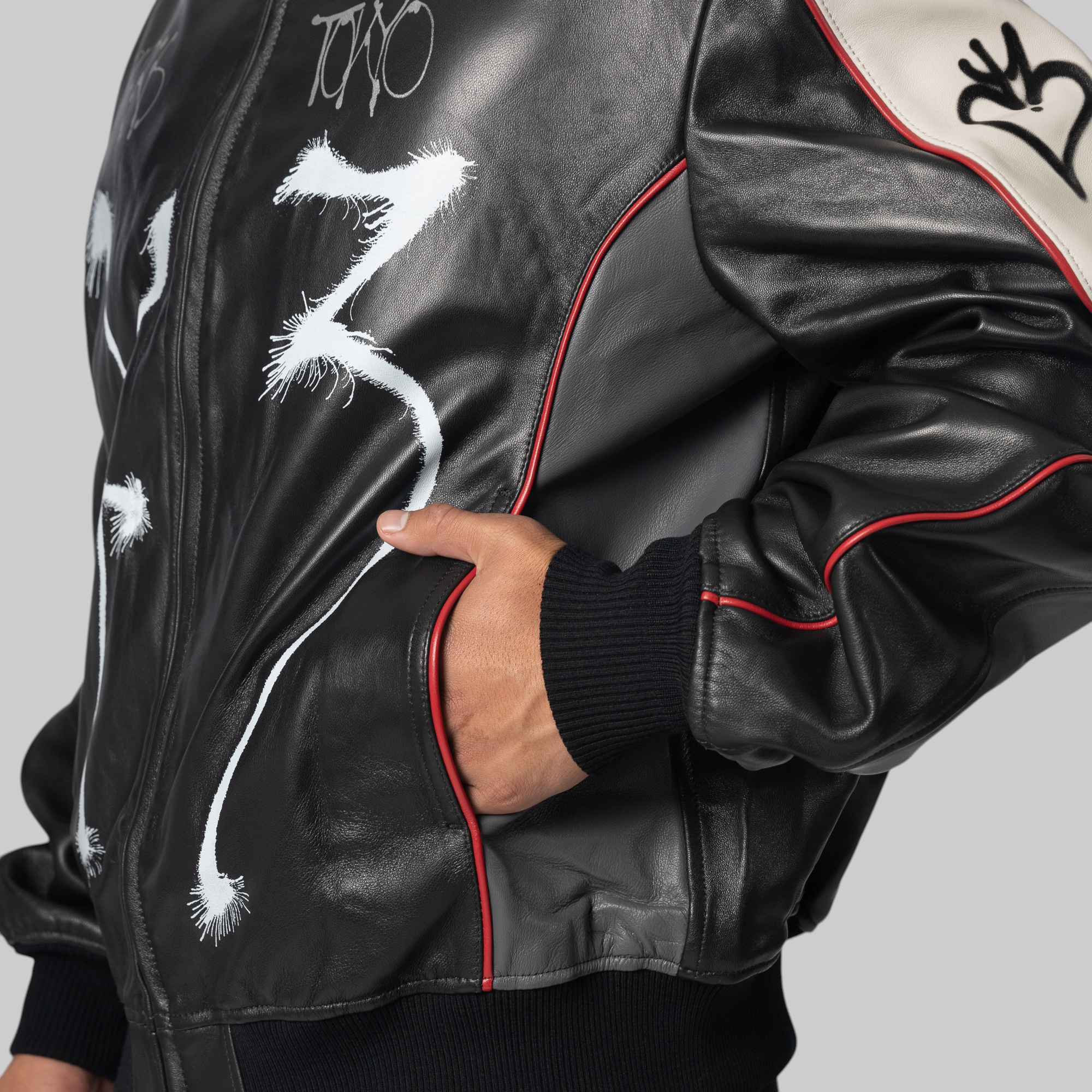 A close-up of a person wearing a black leather bomber jacket with white graffiti-style drawings, red piping, and a beige shoulder panel with a black heart graphic.