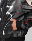 A close-up of a person wearing a black leather bomber jacket with white graffiti-style drawings, red piping, and a beige shoulder panel with a black heart graphic.