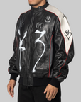 A black leather bomber jacket with white graffiti-style drawings, red piping, and a cream-colored shoulder panel, shown on a model against a solid gray background.