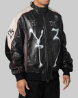 A person wearing a black leather bomber jacket with white graphic text and symbols, cream shoulder panels, and thin red piping, photographed against a plain light gray background.