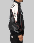 A person shown in side profile wearing a black leather jacket with a large white yoke on the back, red piping, and black graphic prints, paired with black Carhartt pants against a solid gray background.