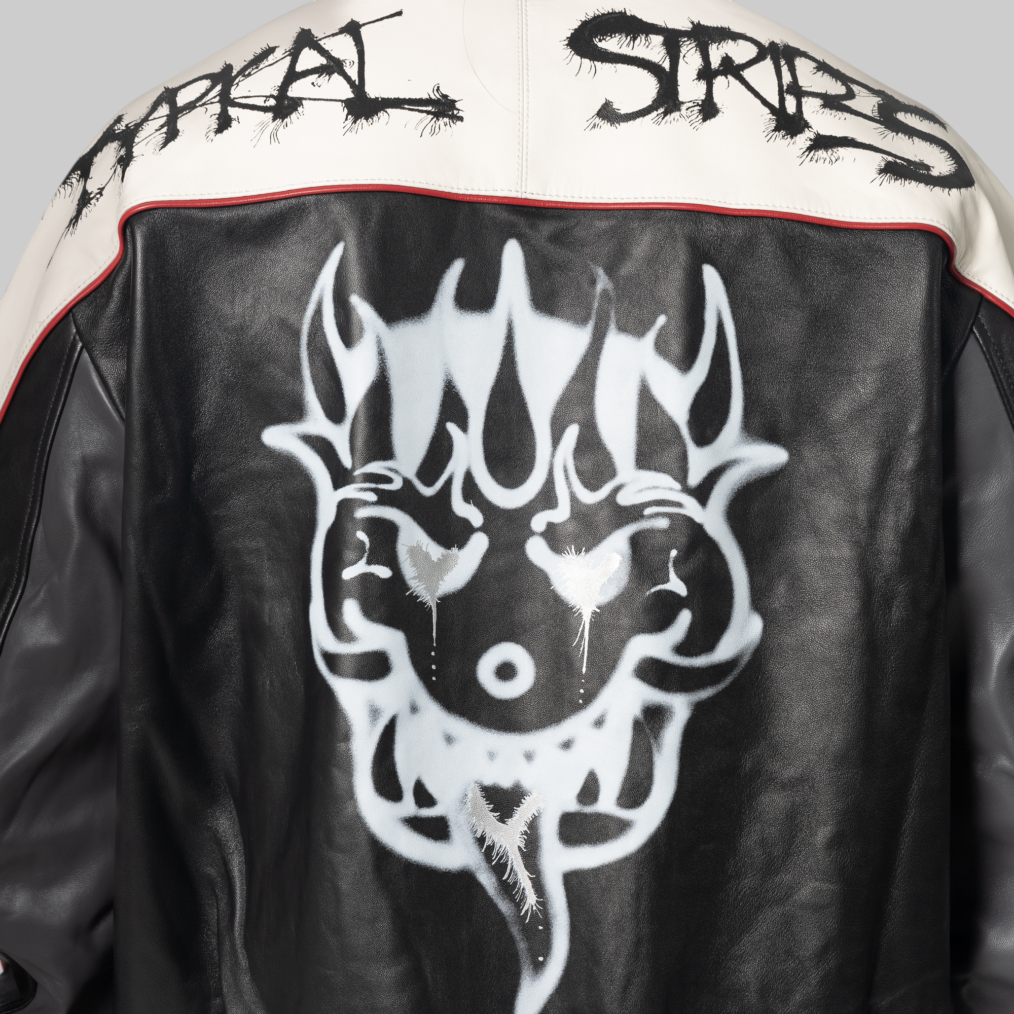 Close-up of the back of a black leather jacket with a large white skull graphic, a cream shoulder yoke with black distressed lettering, and a thin red piping detail.