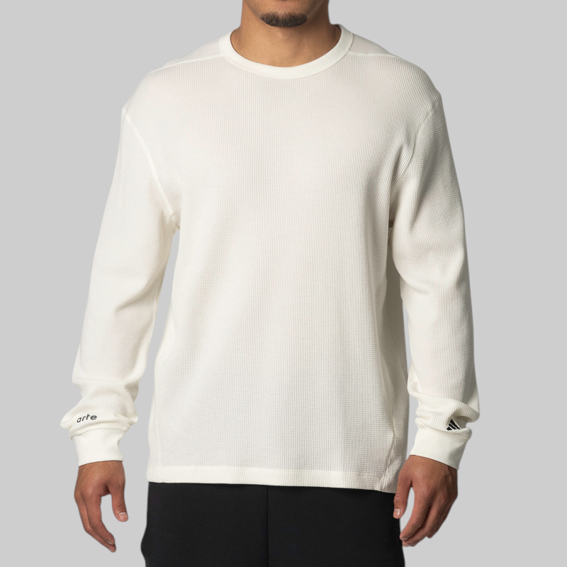 A man wearing a cream-colored, long-sleeve waffle-knit crewneck sweatshirt with small black Adidas branding on the cuffs, paired with black pants, against a solid gray background.