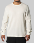 A man wearing a cream-colored, long-sleeve waffle-knit crewneck sweatshirt with small black Adidas branding on the cuffs, paired with black pants, against a solid gray background.