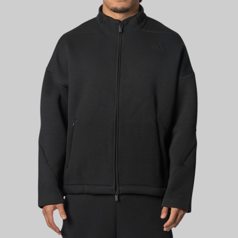 A man wearing a black full-zip athletic jacket with a stand collar, side zip pockets, and a subtle tonal Adidas logo on the left chest, shown against a plain light gray studio background.