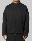 A man wearing a black full-zip athletic jacket with a stand collar, side zip pockets, and a subtle tonal Adidas logo on the left chest, shown against a plain light gray studio background.