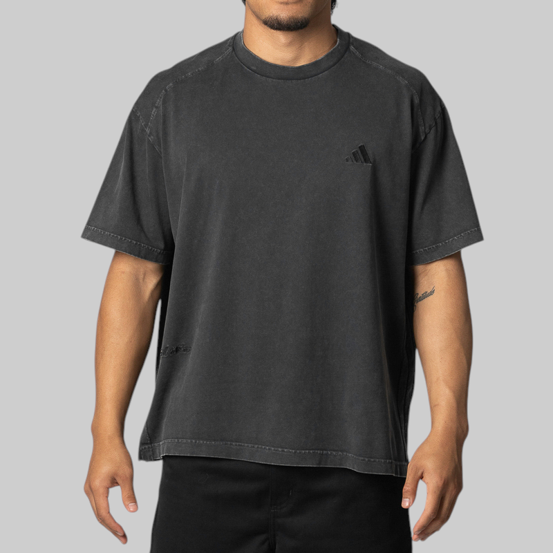A man wearing a faded black short-sleeve t-shirt with a small black embroidered Adidas logo on the left chest, shown against a solid gray background.