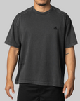 A man wearing a faded black short-sleeve t-shirt with a small black embroidered Adidas logo on the left chest, shown against a solid gray background.