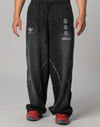 A person wearing black Adidas sweatpants with white graphic logos and speckled details, paired with red sneakers, shown from the waist down against a light gray studio background.