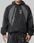 A man wearing a black Adidas track jacket and matching track pants against a solid gray background.