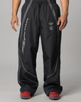 Black Adidas track pants with silver and white graphics, worn by a model standing against a light gray studio background.