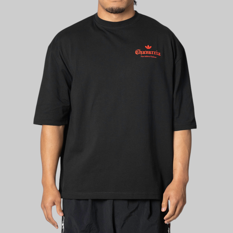 A man wearing an oversized black t-shirt with a red 'Chavarria' logo printed on the left chest, photographed against a solid gray background.