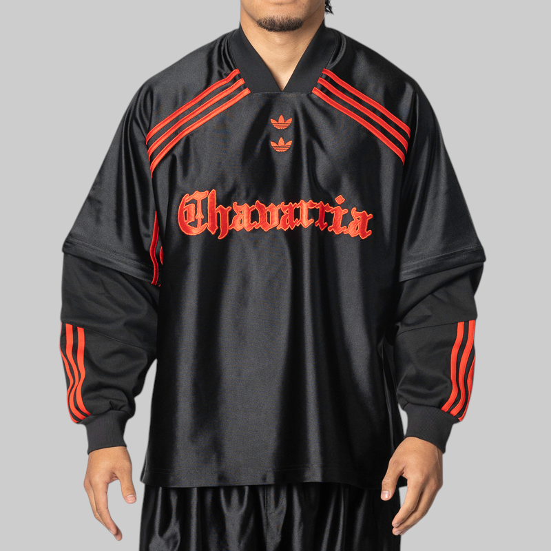 A man wearing a black satin Adidas track jacket with red stripes and 'Chavarria' text on the front, set against a solid gray background.