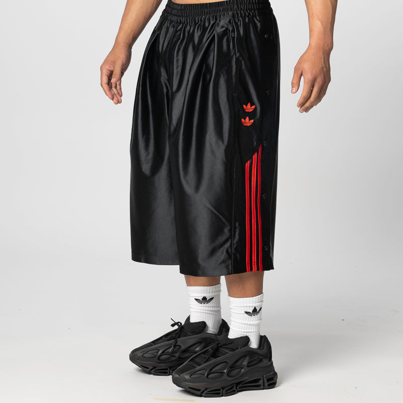 A person wearing black Adidas athletic shorts with red three-stripes and a red trefoil logo, styled with white socks and black sneakers against a light gray studio background.
