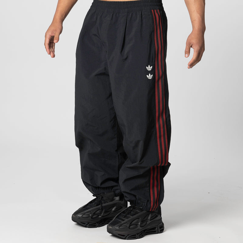 Black track pants with red side stripes and a small white logo, shown on a model from the waist down against a light gray studio background.