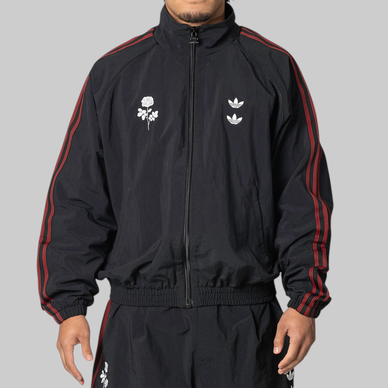 A man wearing a black zip-up track jacket with red stripes down the sleeves and white embroidered chest graphics, paired with matching black track pants, against a solid gray background.