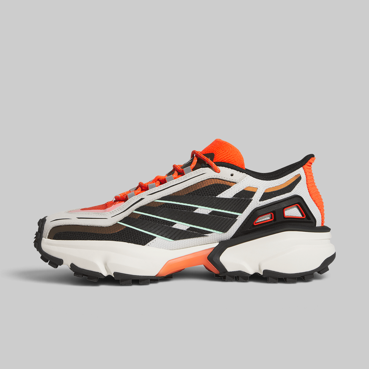 A single athletic sneaker shown in profile against a solid light gray background. The shoe features a white and black mesh upper with bright orange accents on the laces, collar, and midsole, and a chunky white and black sole.