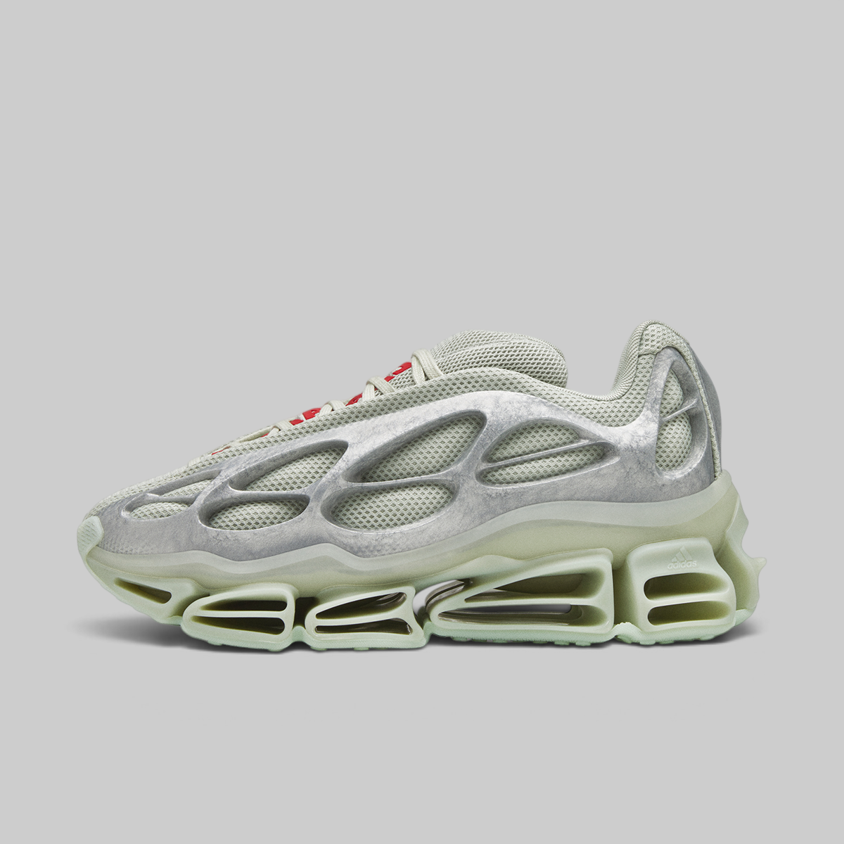 A single futuristic sneaker shown in profile against a solid gray background. The shoe features a metallic silver upper with a textured, organic shell-like overlay, a thick translucent pale green midsole, and small red accents on the laces.