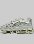 A single futuristic sneaker shown in profile against a solid gray background. The shoe features a metallic silver upper with a textured, organic shell-like overlay, a thick translucent pale green midsole, and small red accents on the laces.