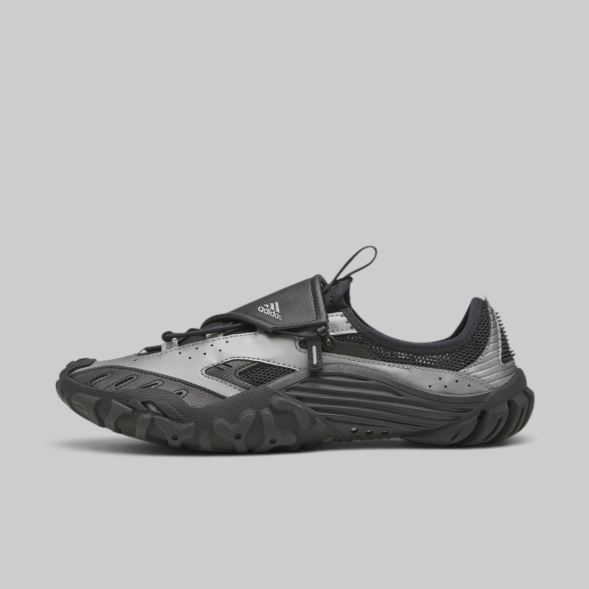 A single Adidas low-top shoe shown in profile against a solid gray background. The shoe features a metallic silver and black upper with a prominent black hook-and-loop strap across the instep, a black textured sole, and the white Adidas logo on the strap.
