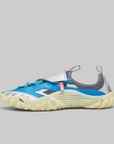 A single athletic sneaker shown in profile against a solid gray background. The shoe features a bright blue upper, a white strap with a silver buckle, a gray mesh collar, and a thick, textured cream-colored sole.
