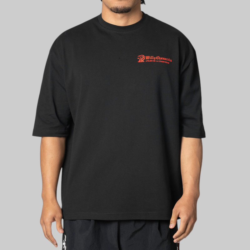 A man wearing an oversized black t-shirt with a red 'Willy Chavarria' logo on the left chest, photographed against a solid gray background.