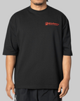 A man wearing an oversized black t-shirt with a red 'Willy Chavarria' logo on the left chest, photographed against a solid gray background.