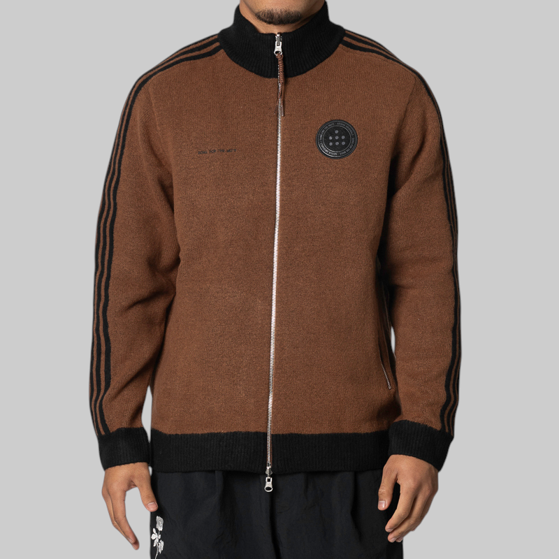 A man wearing a brown knit zip-up track jacket with black ribbed trim, black shoulder stripes, and a circular black chest patch, photographed against a plain light gray studio background.