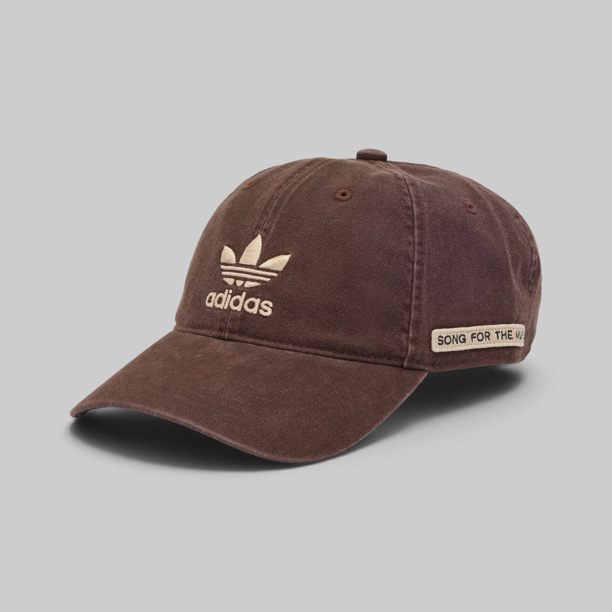 A brown washed cotton baseball cap featuring a beige embroidered Adidas trefoil logo and brand name on the front, and a beige patch on the side that reads 'SONG FOR THE NOY'. The cap is photographed on a neutral gray background.