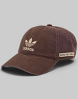 A brown washed cotton baseball cap featuring a beige embroidered Adidas trefoil logo and brand name on the front, and a beige patch on the side that reads 'SONG FOR THE NOY'. The cap is photographed on a neutral gray background.