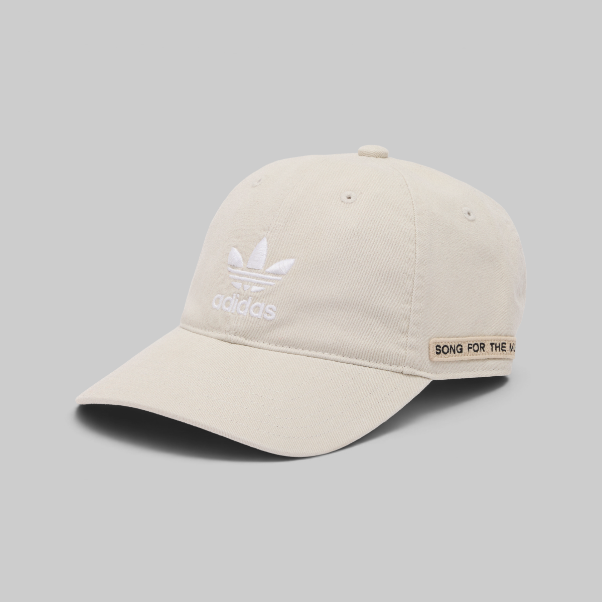 A light beige baseball cap by adidas, featuring a white embroidered trefoil logo and 'adidas' text on the front, and a small tan patch on the side with the text 'SONG FOR THE MAJESTY OF ALL CREATION'. The cap is photographed on a neutral gray background.