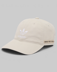 A light beige baseball cap by adidas, featuring a white embroidered trefoil logo and 'adidas' text on the front, and a small tan patch on the side with the text 'SONG FOR THE MAJESTY OF ALL CREATION'. The cap is photographed on a neutral gray background.