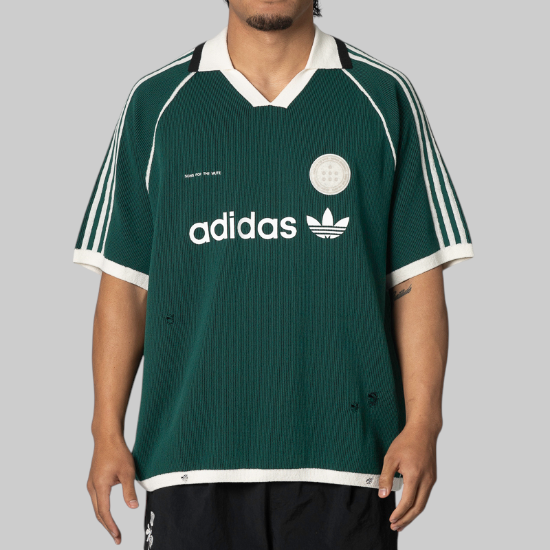 A man wearing a dark green Adidas short-sleeve soccer jersey with white stripes, a white collar, and white logo details, shown against a solid gray background.
