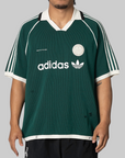 A man wearing a dark green Adidas short-sleeve soccer jersey with white stripes, a white collar, and white logo details, shown against a solid gray background.