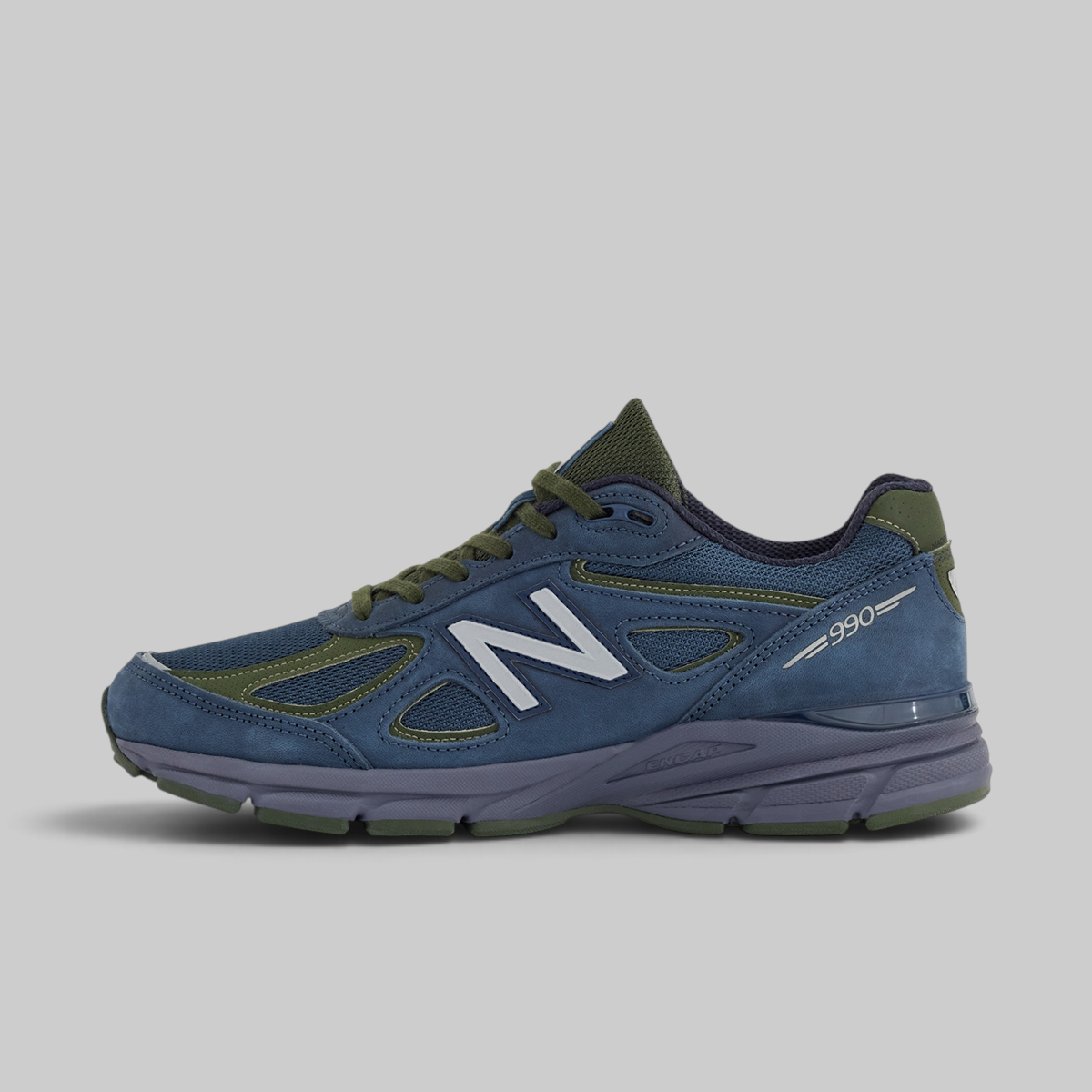 A New Balance 990 sneaker in blue suede and mesh with olive green accents and a white 'N' logo, shown from a side profile on a light gray background.