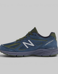 A New Balance 990 sneaker in blue suede and mesh with olive green accents and a white 'N' logo, shown from a side profile on a light gray background.