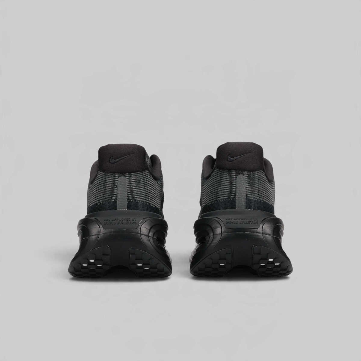 A pair of Nike sneakers shown from the back, featuring dark gray and black mesh and synthetic materials with chunky black soles, set against a solid light gray background.