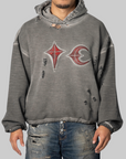 A man wearing a distressed gray hooded sweatshirt with a red and white graphic logo on the chest, paired with ripped blue jeans, against a solid gray background.