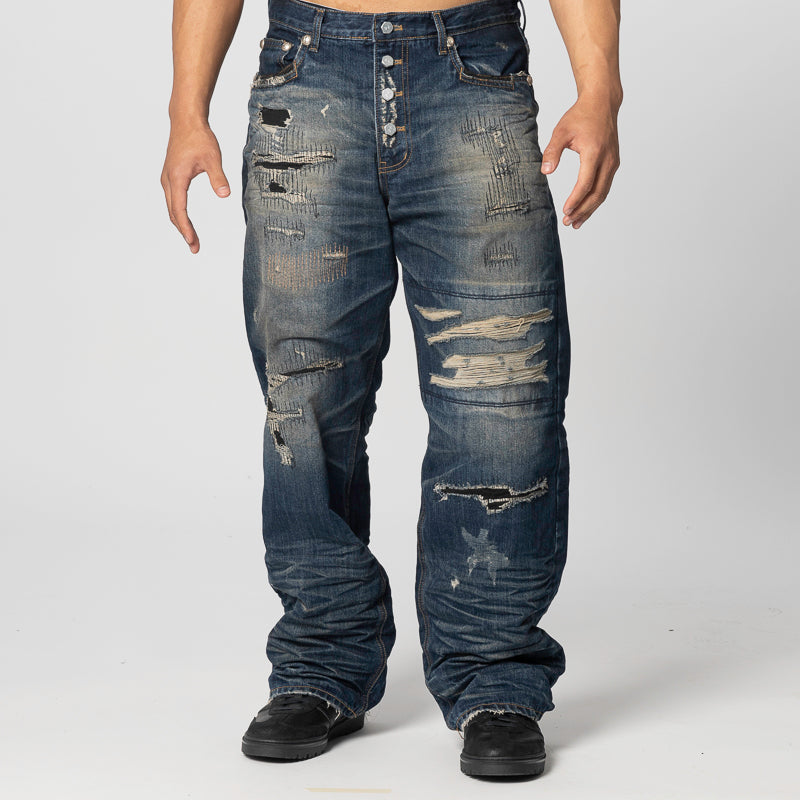 A person modeling a pair of heavily distressed blue denim jeans with ripped details and frayed edges, paired with black shoes, against a light gray studio background.