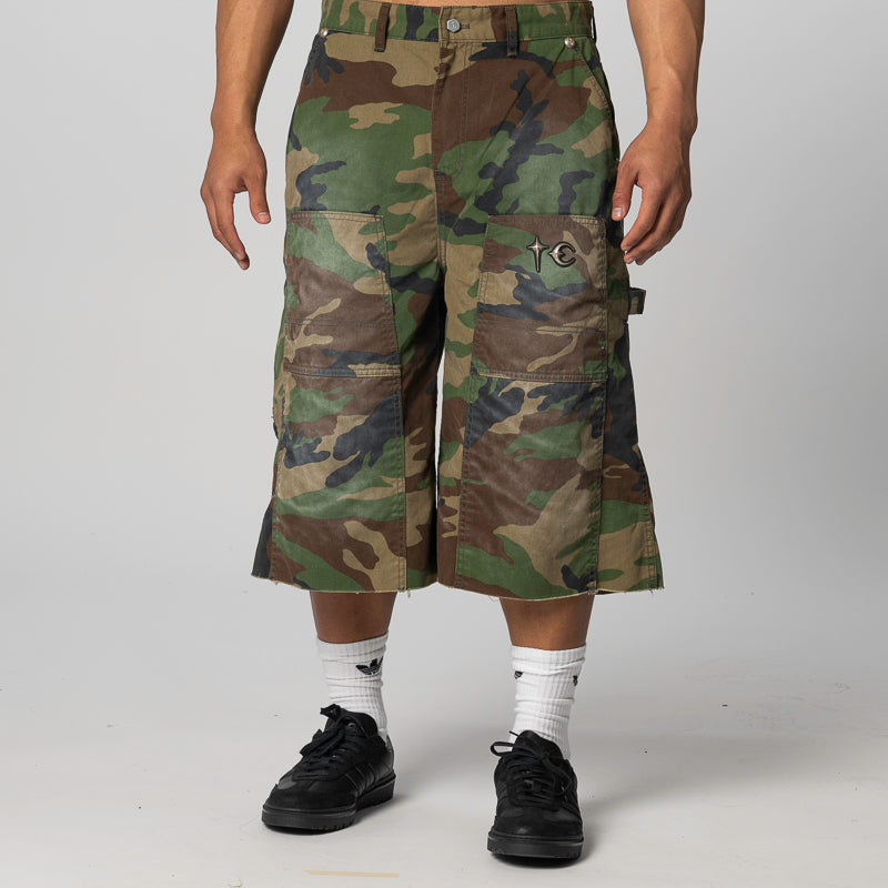 A person wearing knee-length camouflage cargo shorts with large front flap pockets, shown from the waist down against a light gray studio background. The shorts feature a woodland camo pattern in green, brown, black, and tan, with a small embroidered chest logo.