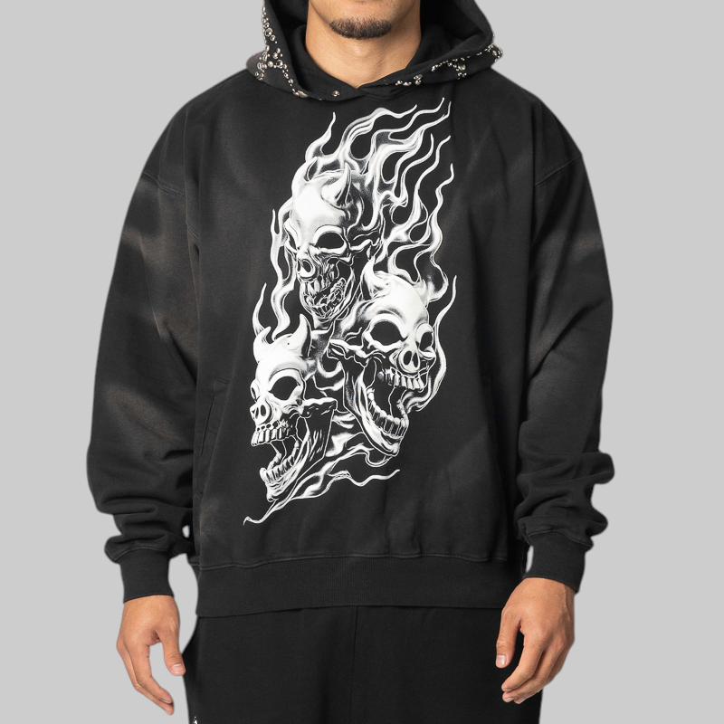 A man wearing a black pullover hoodie with a large white graphic print of three skulls and flames on the front, shown against a solid gray background.