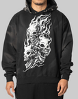 A man wearing a black pullover hoodie with a large white graphic print of three skulls and flames on the front, shown against a solid gray background.