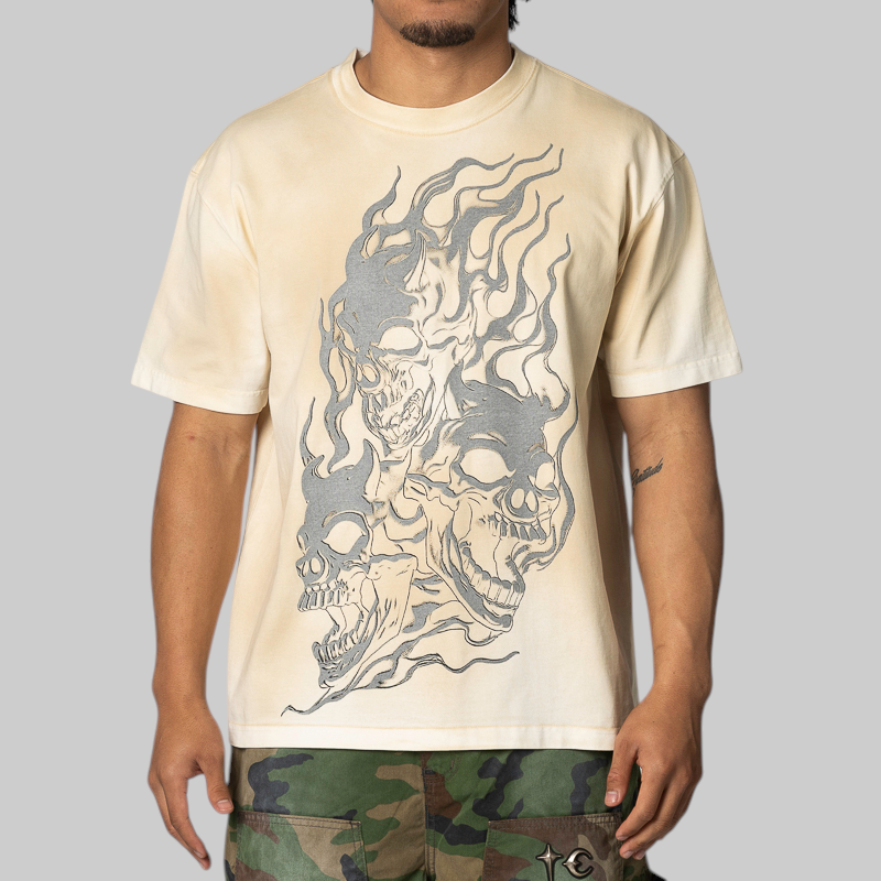 A man wearing a cream-colored t-shirt with a large gray graphic of three skulls in flames, paired with camouflage pants, against a solid gray background.