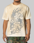 A man wearing a cream-colored t-shirt with a large gray graphic of three skulls in flames, paired with camouflage pants, against a solid gray background.