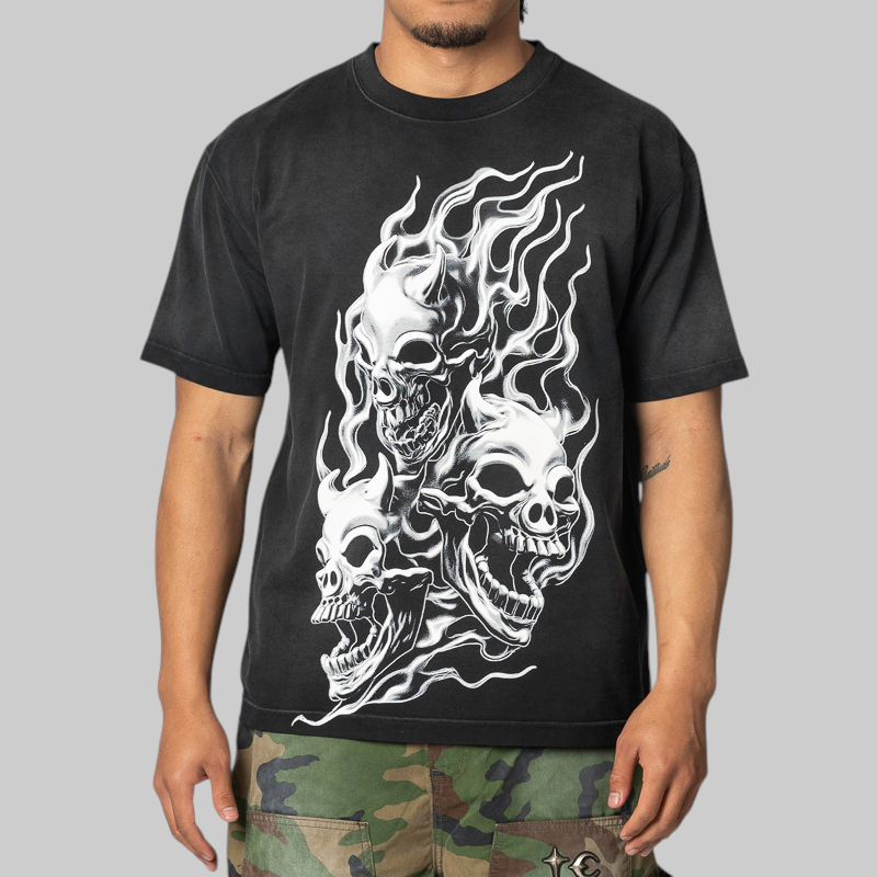 A man wearing a black short-sleeve t-shirt with a large white graphic of three skulls surrounded by flames, paired with camouflage pants, against a solid gray background.